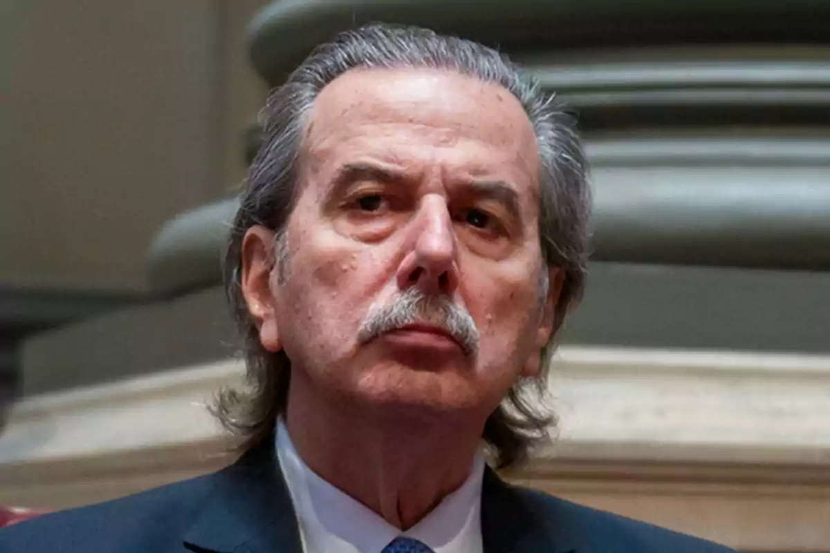 Hombre de cabello canoso y bigote con expresión seria vistiendo traje oscuro y corbata azul en un entorno institucional