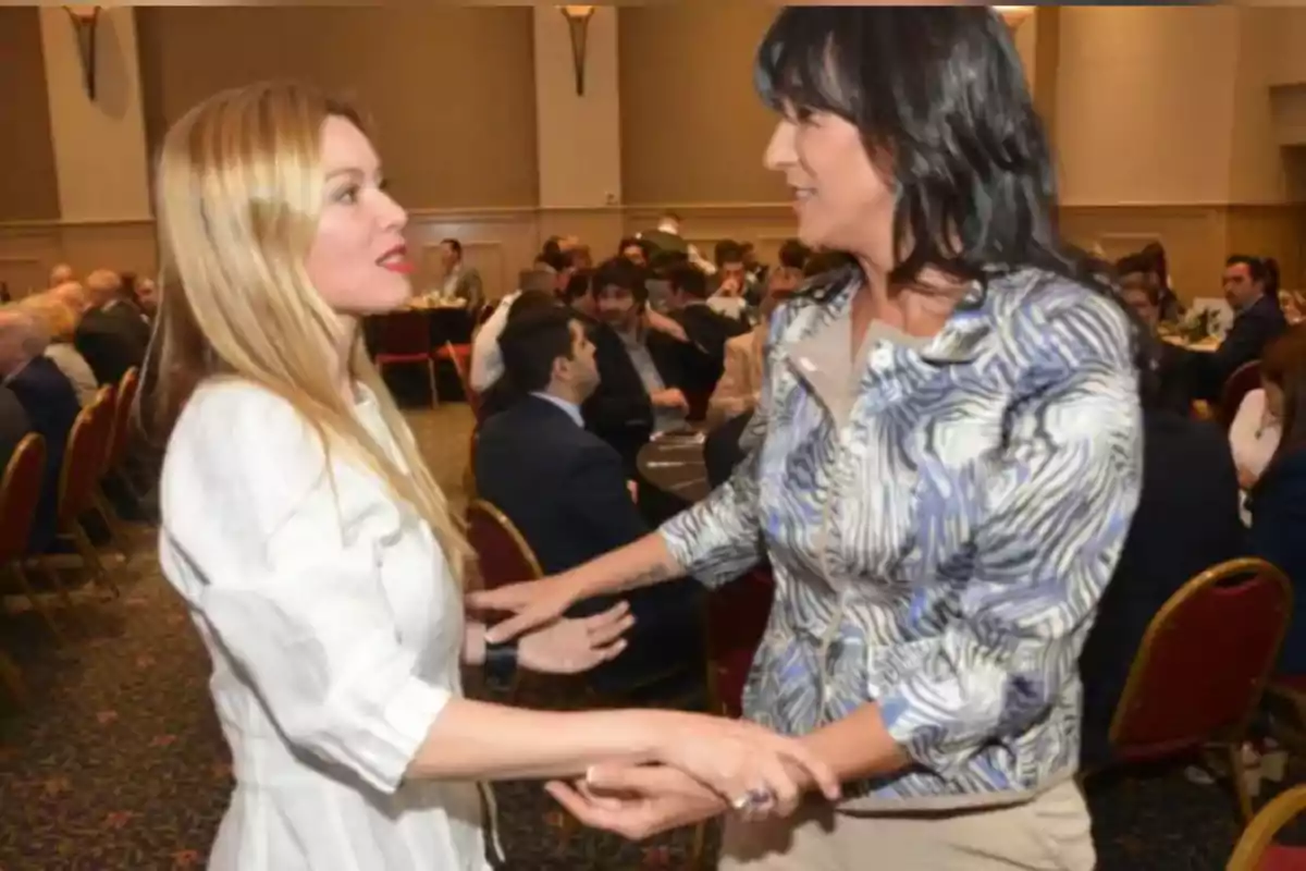 Dos mujeres se saludan de pie en un salón lleno de personas sentadas en sillas rojas durante un evento formal Dos mujeres se saludan de pie en un salón lleno de personas sentadas en sillas rojas durante un evento formal