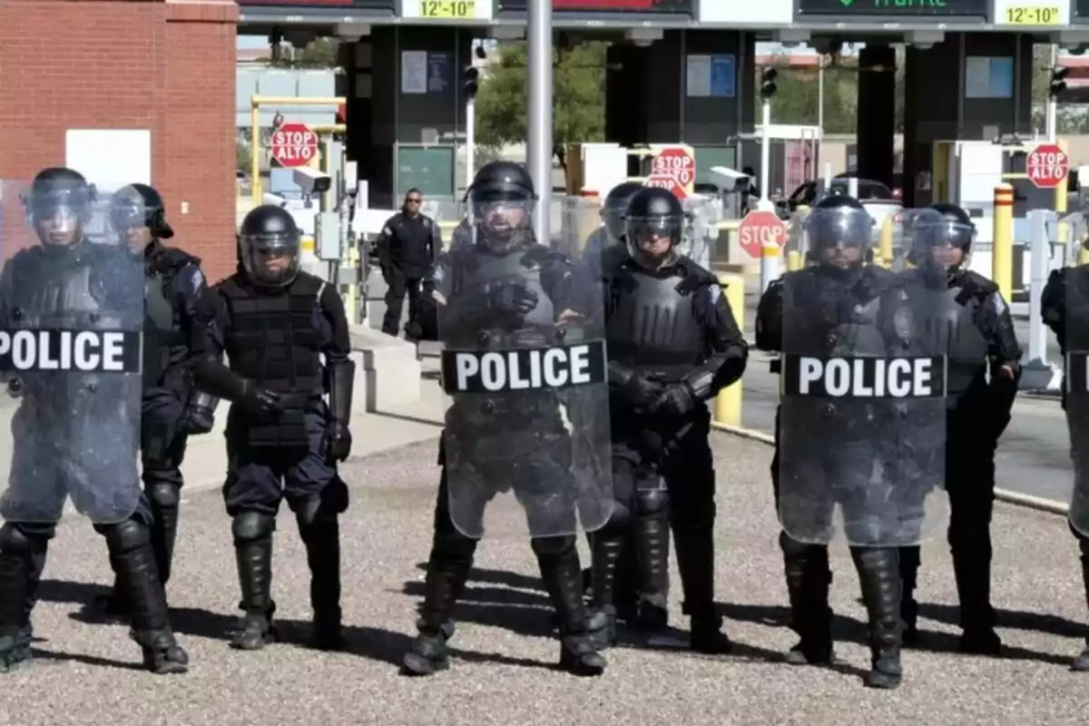 Un grupo de policías antidisturbios con equipo de protección y escudos se encuentra alineado frente a una entrada con señales de alto.