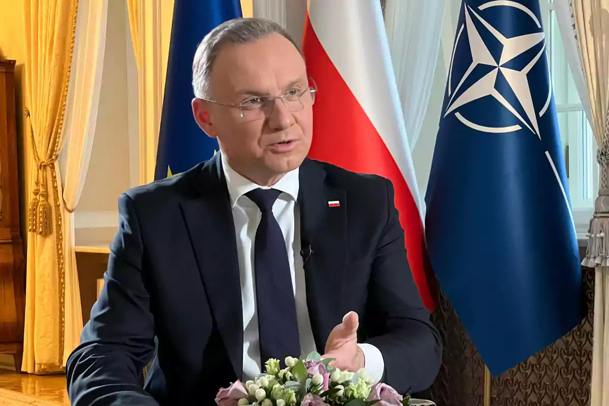 Un hombre en traje oscuro está sentado frente a banderas de la Unión Europea, Polonia y la OTAN, hablando y gesticulando con la mano.