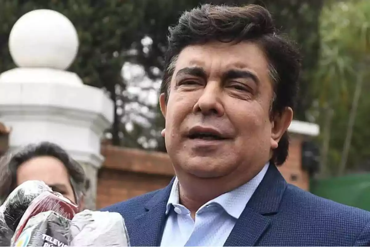 Un hombre de mediana edad con cabello oscuro y traje azul está hablando al aire libre, con un micrófono y una cámara en primer plano.