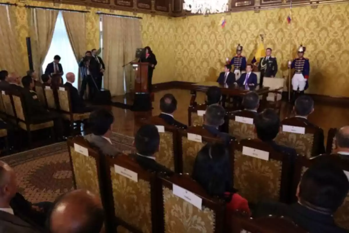 A conference room with people seated, a podium with a person speaking, and uniformed guards standing in the back.