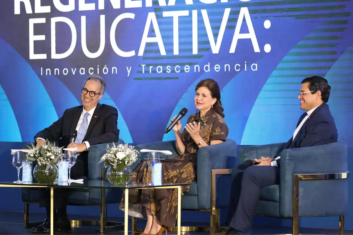 Tres personas sentadas en sillones azules conversan en un panel sobre regeneración educativa, con un fondo azul y flores blancas sobre la mesa frente a ellos