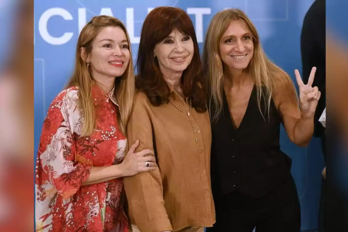 Tres mujeres posan sonrientes frente a un fondo azul, una de ellas hace el gesto de la victoria con la mano Tres mujeres posan sonrientes frente a un fondo azul, una de ellas hace el gesto de la victoria con la mano