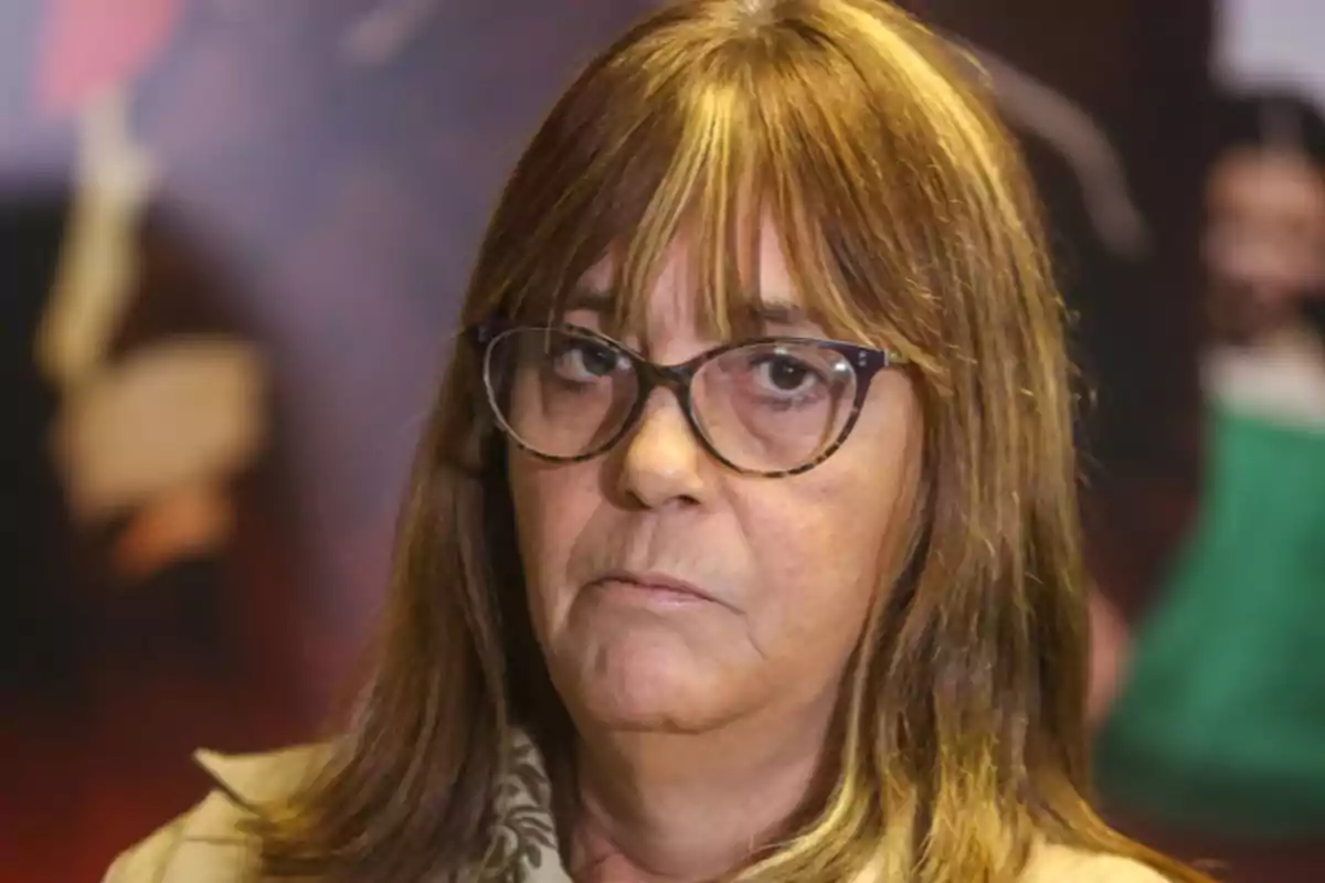 A woman with glasses and light brown hair looks at the camera with a serious expression and a blurred background. A woman with glasses and light brown hair looks at the camera with a serious expression and a blurred background.