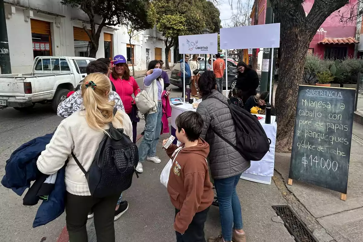 Un grupo de personas se reúne alrededor de un puesto en la calle, donde se ofrece información y hay un cartel con un menú que incluye milanesa a caballo y panqueques. Un grupo de personas se reúne alrededor de un puesto en la calle, donde se ofrece información y hay un cartel con un menú que incluye milanesa a caballo y panqueques.