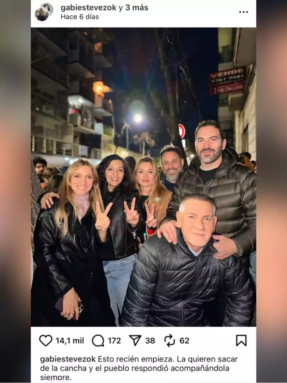 Grupo de personas posando sonrientes en una calle de noche, algunas haciendo el gesto de la victoria con los dedos, con edificios y luces de fondo Grupo de personas posando sonrientes en una calle de noche, algunas haciendo el gesto de la victoria con los dedos, con edificios y luces de fondo