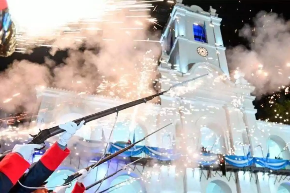 People in uniforms firing salutes in front of a building illuminated with blue and white lights and decorations.