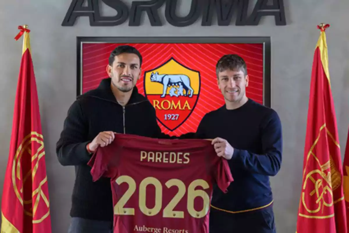 Dos personas sostienen una camiseta de fútbol con el nombre "Paredes" y el número 2026 frente a un escudo del equipo AS Roma.