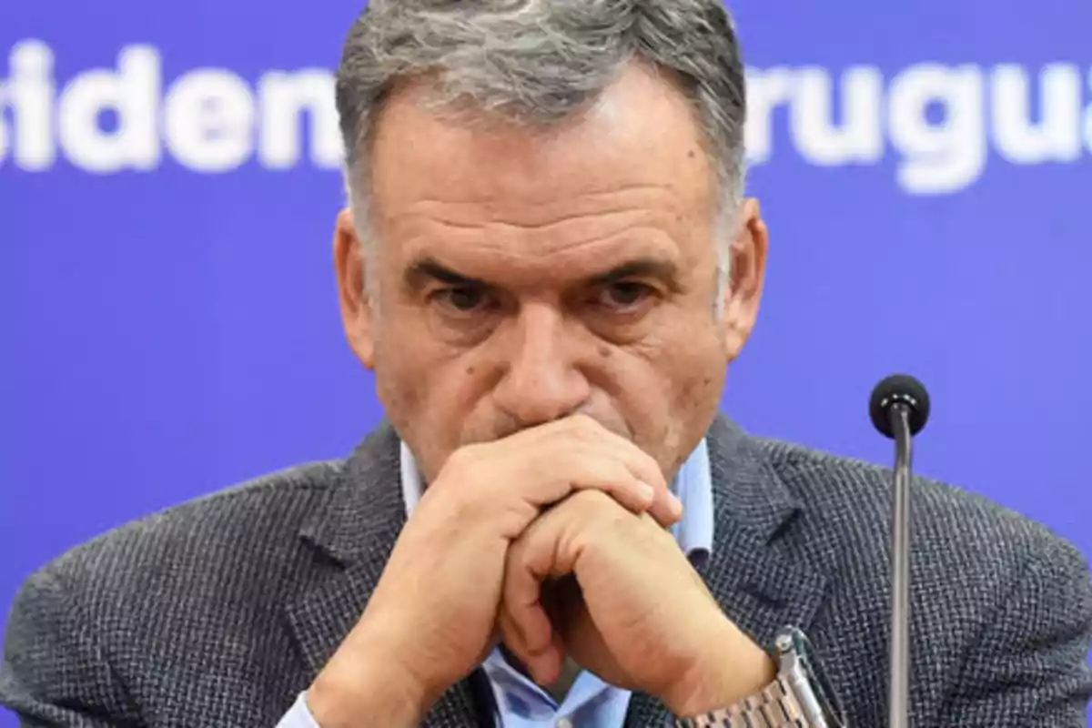 Hombre de cabello canoso con expresión seria y manos entrelazadas frente a su rostro sentado frente a un micrófono con fondo azul Hombre de cabello canoso con expresión seria y manos entrelazadas frente a su rostro sentado frente a un micrófono con fondo azul
