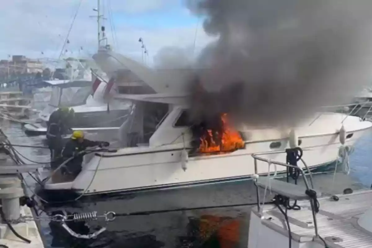 A white yacht at a dock is on fire while two firefighters try to put out the flames and a large cloud of black smoke rises into the sky.