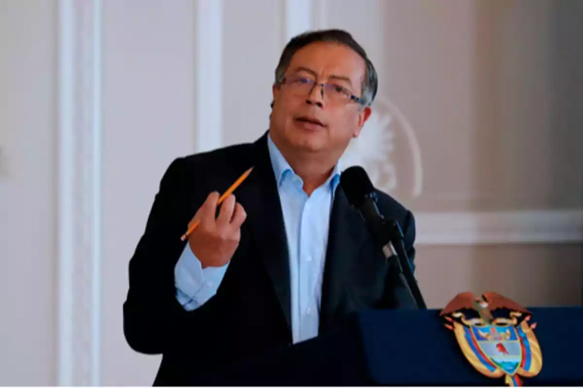 Un hombre con gafas y traje oscuro habla en un podio con un escudo, sosteniendo un lápiz en la mano.