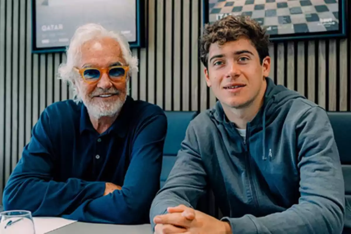 Two men sitting together at a table, one older with a white beard and colorful glasses and the other younger wearing a gray sweatjersey, both smiling at the camera. Two men sitting together at a table, one older with a white beard and colorful glasses and the other younger wearing a gray sweatjersey, both smiling at the camera.