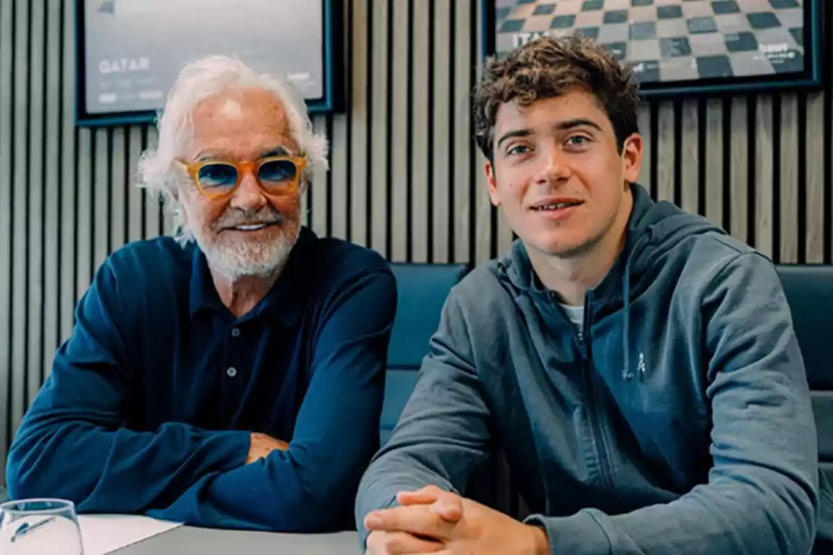 Two people sitting at a table, one of them is older with a beard and sunglasses, the other is younger and wearing a hoodie. Two people sitting at a table, one of them is older with a beard and sunglasses, the other is younger and wearing a hoodie.