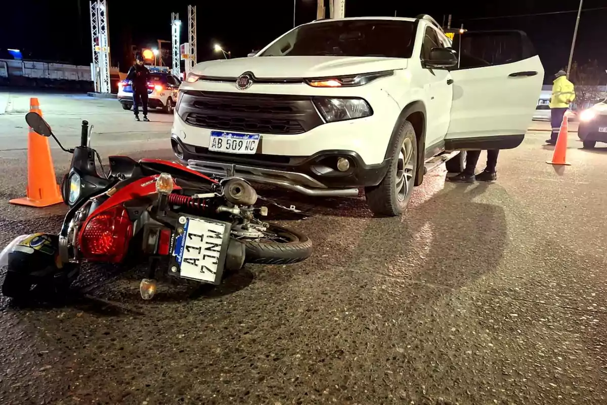 Una motocicleta caída en el suelo frente a una camioneta blanca con la puerta abierta en una calle de noche, rodeados de conos naranjas y presencia policial