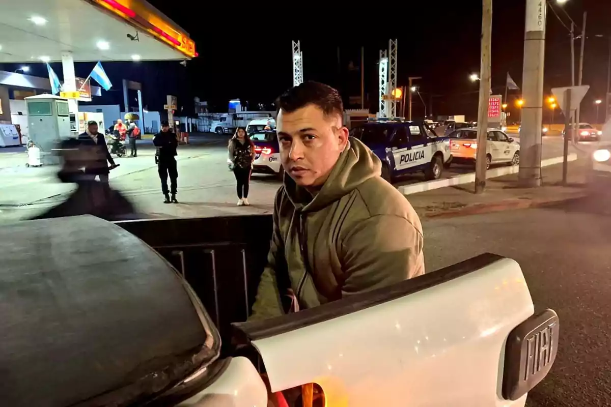Man in a green jacket sitting in the back of a white pickup truck at a gas station at night with police cars and several people in the background Man in a green jacket sitting in the back of a white pickup truck at a gas station at night with police cars and several people in the background