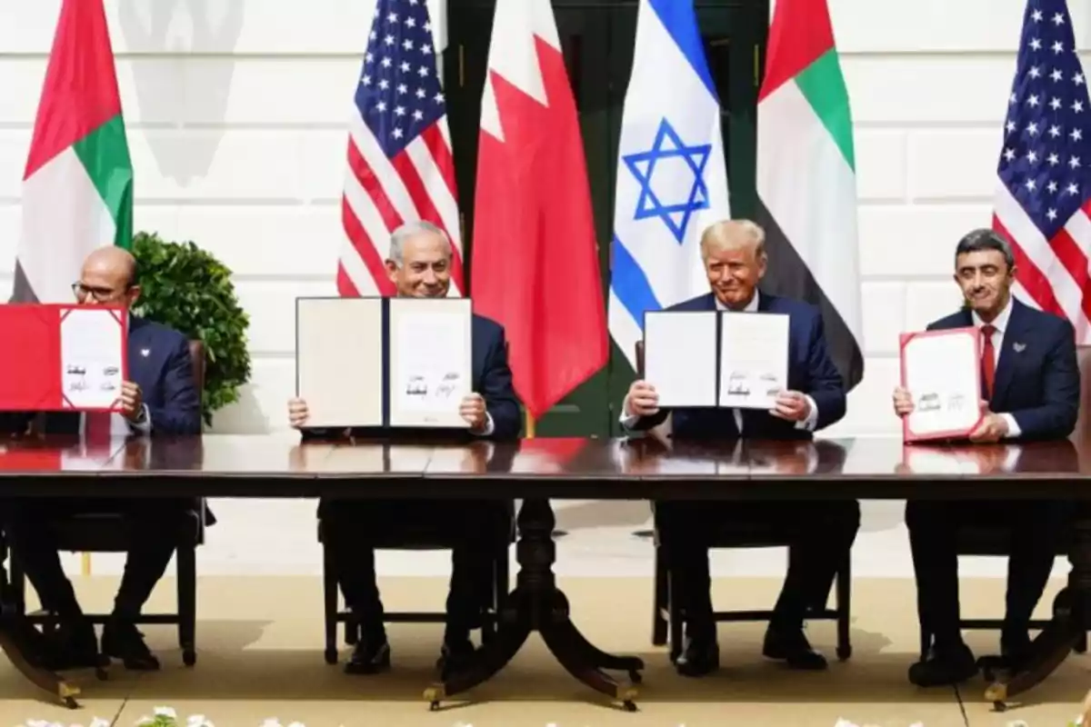 Cuatro personas sentadas en una mesa sosteniendo documentos firmados, con banderas de varios países detrás de ellos. Cuatro personas sentadas en una mesa sosteniendo documentos firmados, con banderas de varios países detrás de ellos.