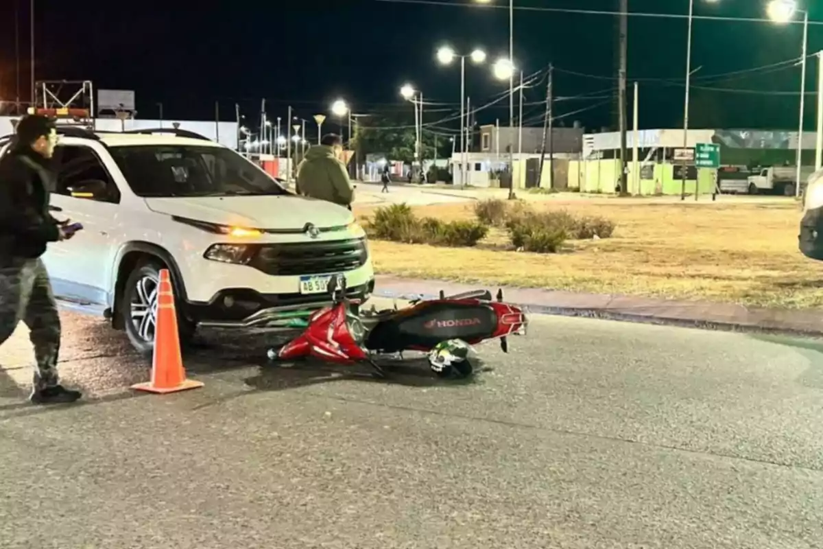 A white car and a red motorcycle lying on the ground after an accident on a well-lit street at night, with people around and an orange cone marking the area. A white car and a red motorcycle lying on the ground after an accident on a well-lit street at night, with people around and an orange cone marking the area.