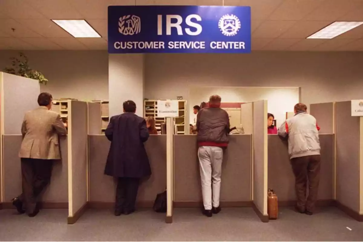 Personas atendiendo en un centro de servicio al cliente del IRS con cubículos y un letrero azul en la parte superior. Personas atendiendo en un centro de servicio al cliente del IRS con cubículos y un letrero azul en la parte superior.