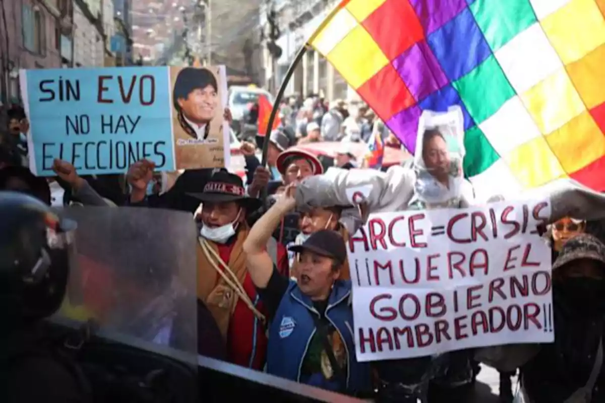 Protesters on a street hold signs and a multicolored flag during a protest in Bolivia.