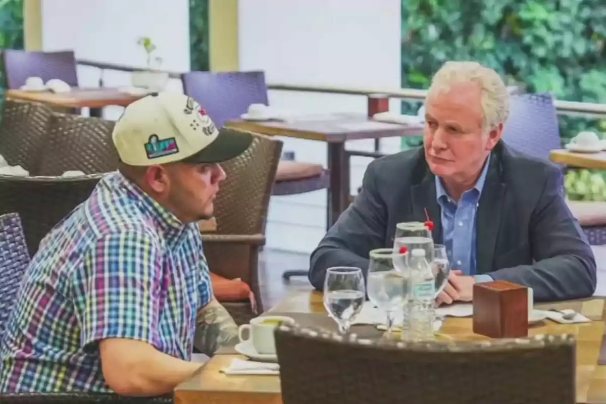 Dos hombres sentados en una mesa de restaurante conversan seriamente rodeados de copas y platos Dos hombres sentados en una mesa de restaurante conversan seriamente rodeados de copas y platos