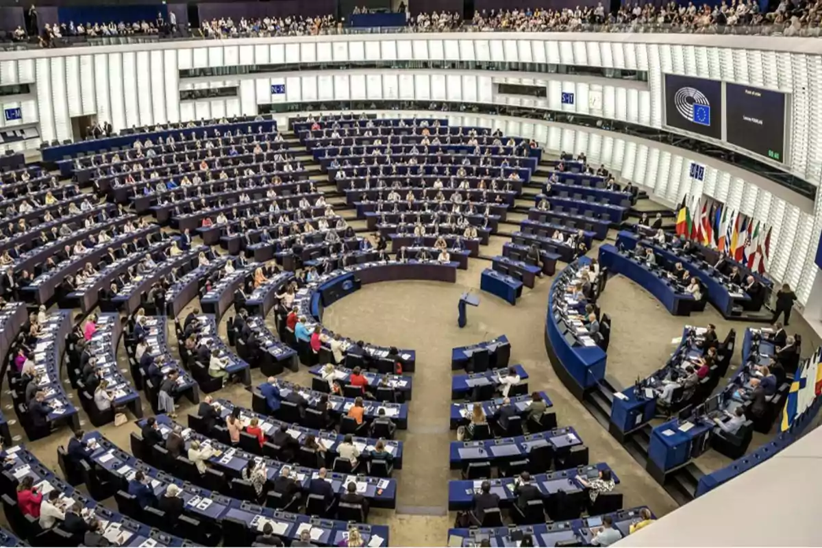 Vista del interior de un parlamento con numerosos asientos ocupados por personas y varias banderas en un lateral.