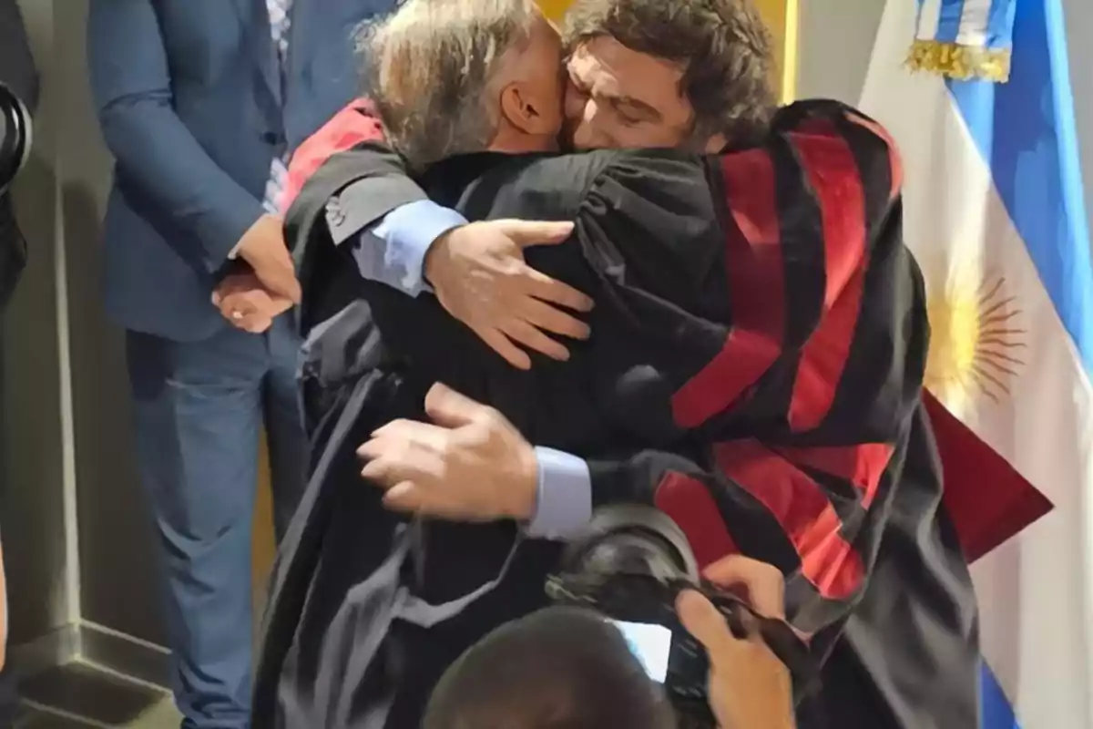 Two people in academic gowns embrace in front of an Argentine flag while a photographer captures the moment. Two people in academic gowns embrace in front of an Argentine flag while a photographer captures the moment.