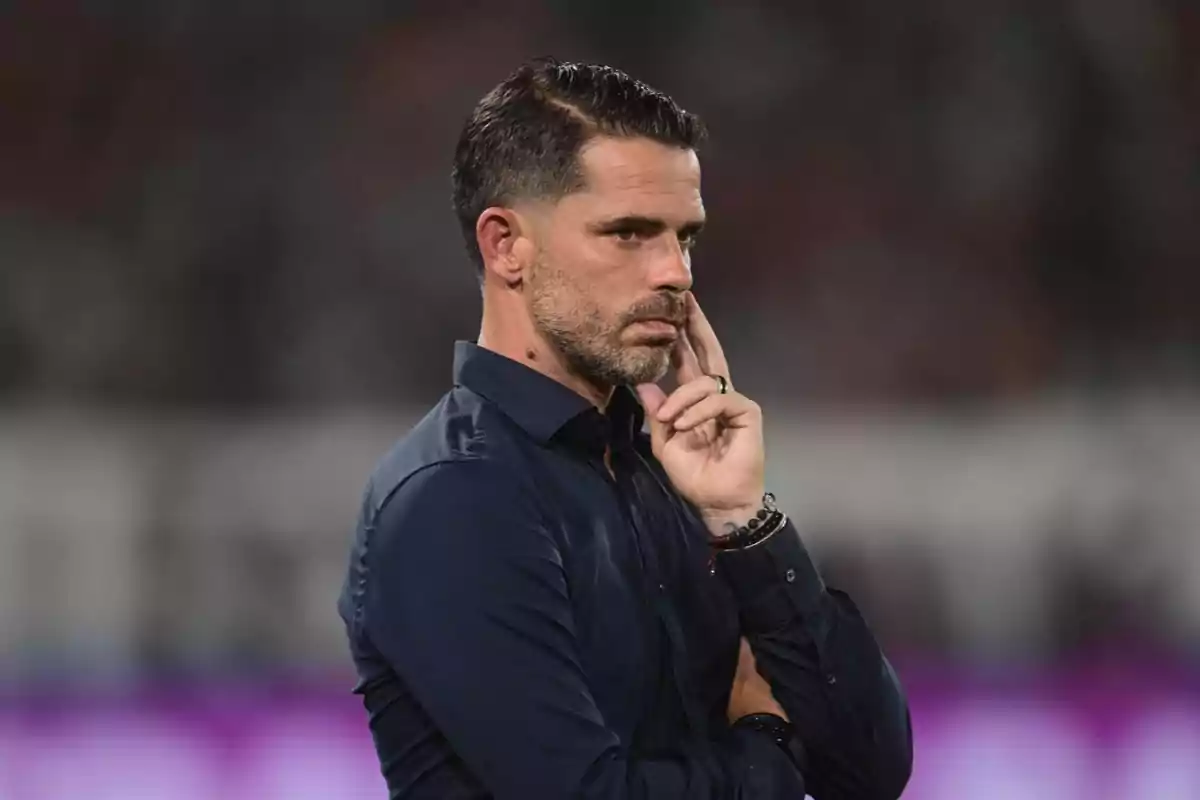 Hombre de cabello corto y barba con camisa azul oscuro en actitud pensativa durante un evento deportivo Hombre de cabello corto y barba con camisa azul oscuro en actitud pensativa durante un evento deportivo