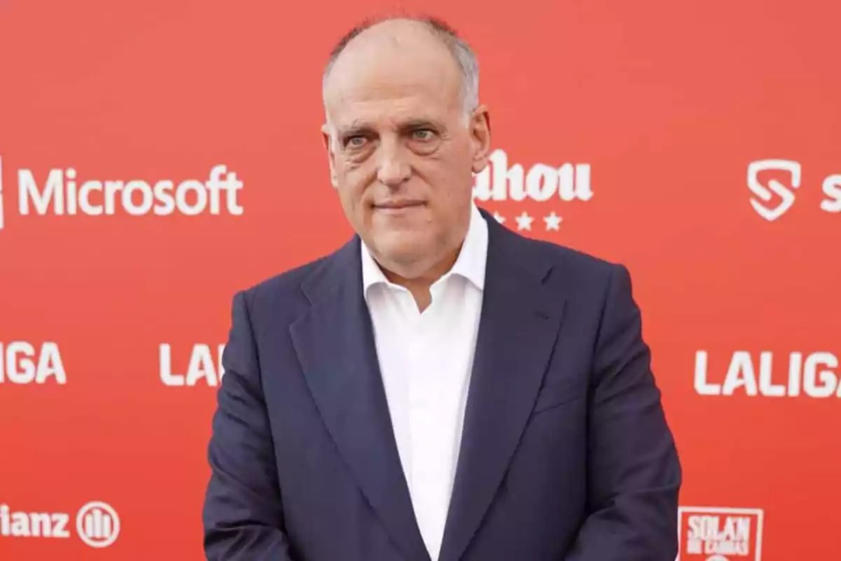 Hombre de mediana edad con traje oscuro y camisa blanca posando frente a un fondo rojo con logotipos de marcas y LaLiga Hombre de mediana edad con traje oscuro y camisa blanca posando frente a un fondo rojo con logotipos de marcas y LaLiga