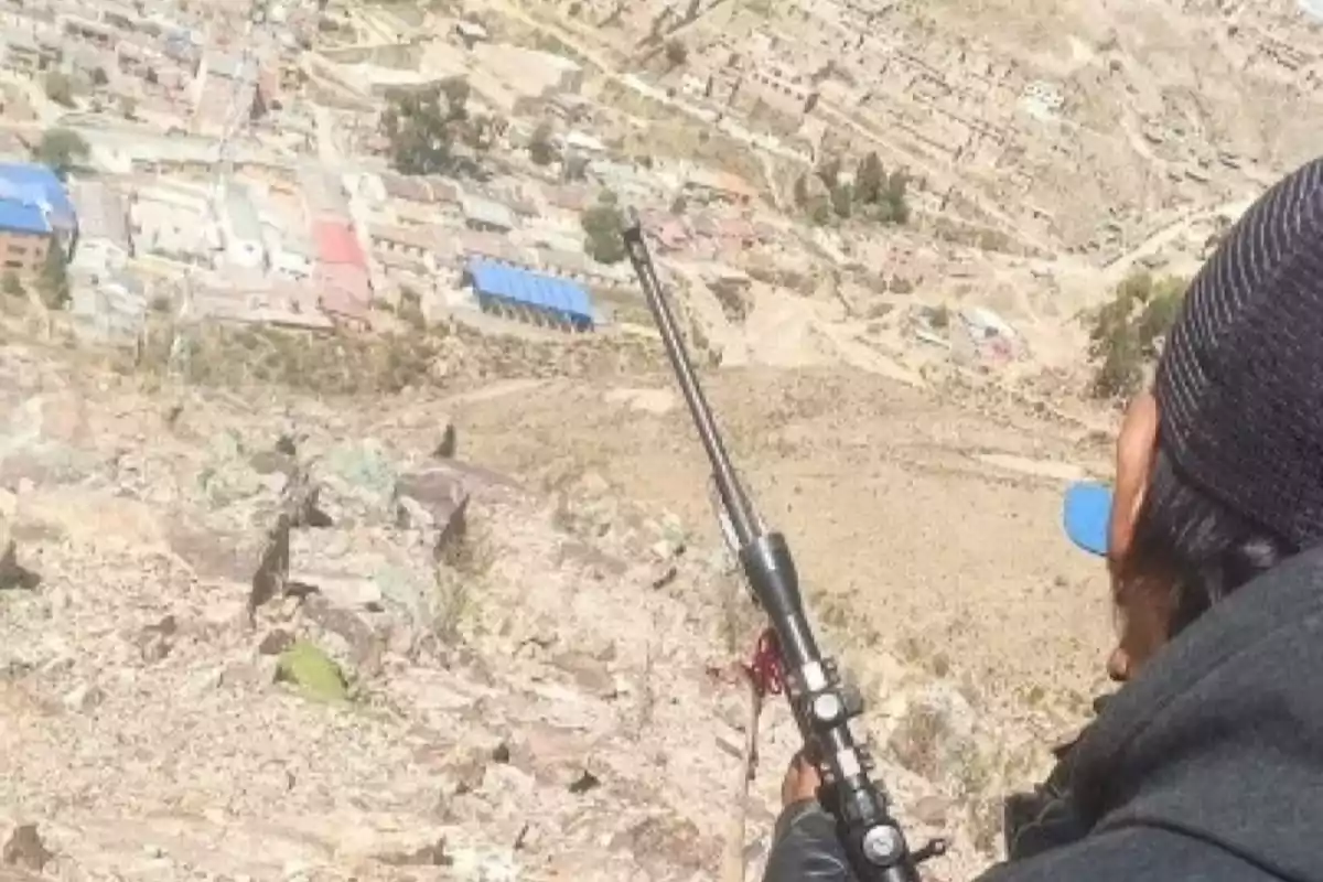 Person wearing a cap holding a rifle in a mountainous area with a village in the background Person wearing a cap holding a rifle in a mountainous area with a village in the background
