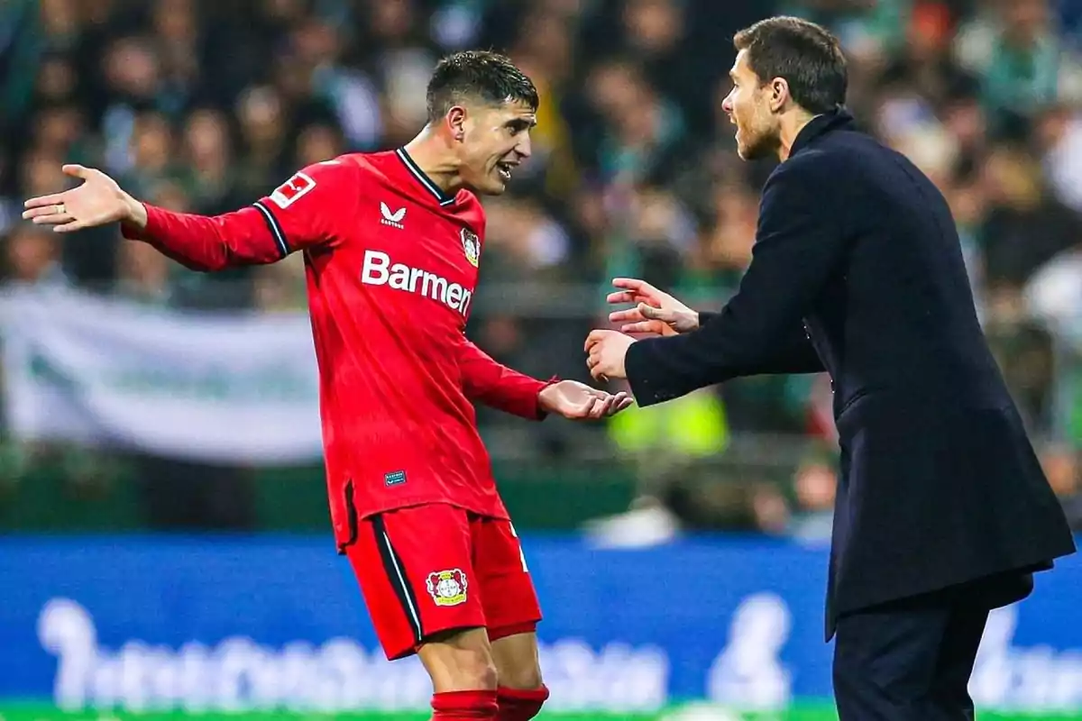 A player in a red uniform gestures with open arms while talking to a man in a suit on a playing field. A player in a red uniform gestures with open arms while talking to a man in a suit on a playing field.