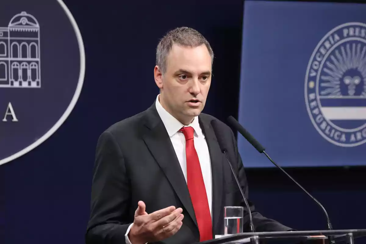 Un hombre con traje y corbata roja hablando en un podio con micrófonos y un fondo con logotipos oficiales.