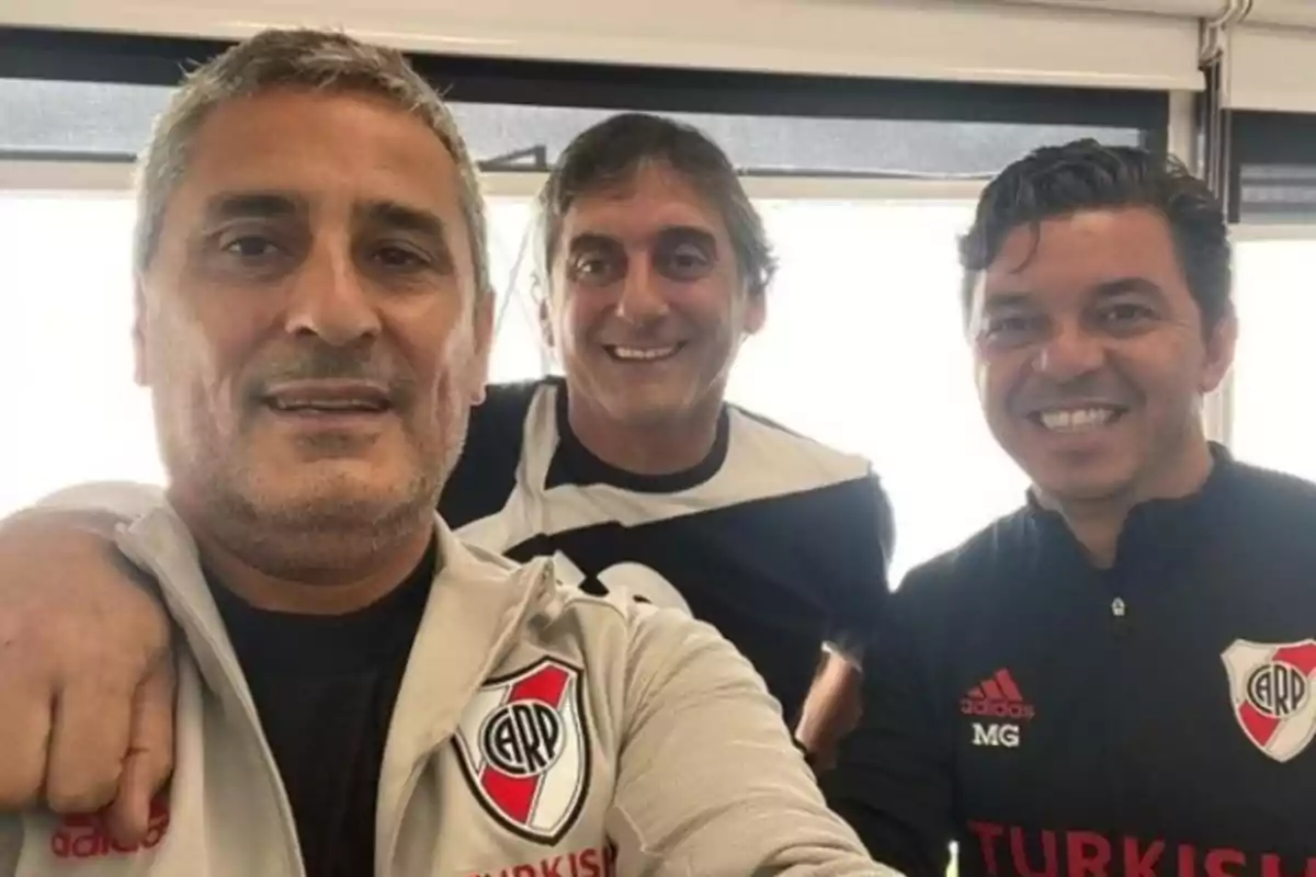 Three men smile at the camera, two of them wearing jackets with a soccer team crest. Three men smile at the camera, two of them wearing jackets with a soccer team crest.