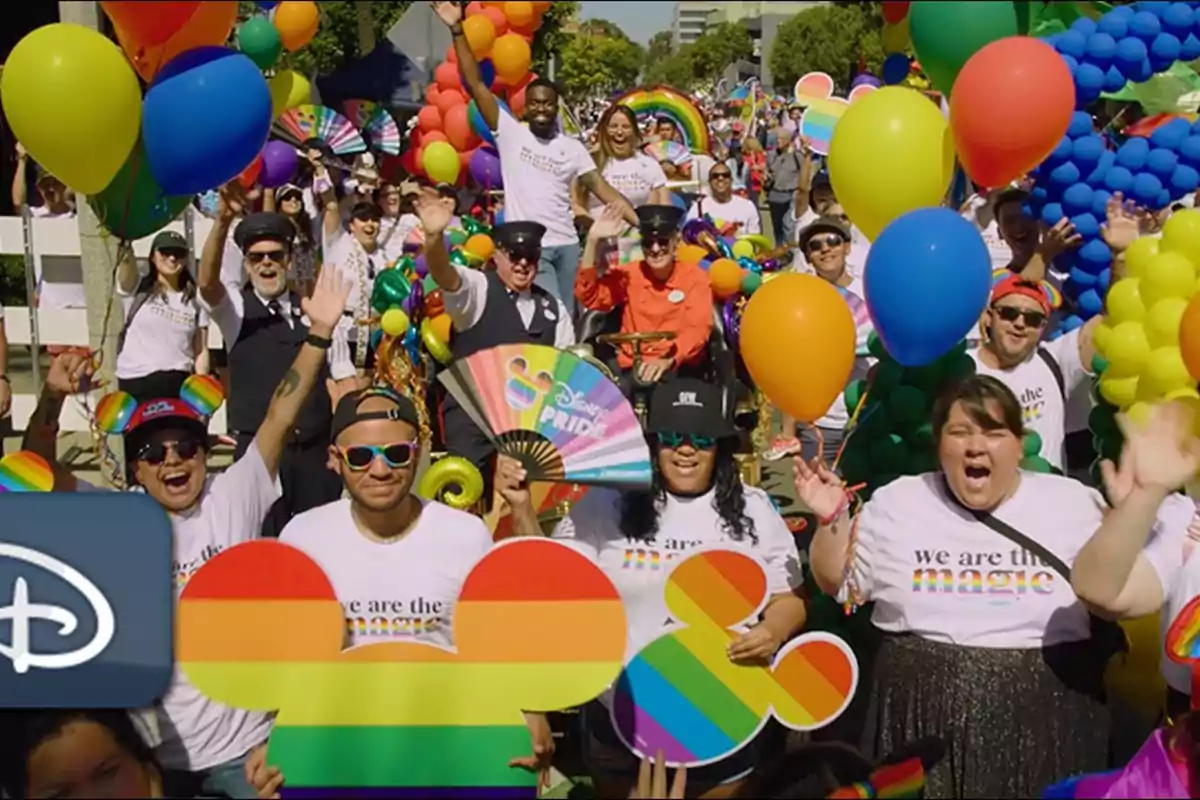 Un grupo de personas celebra en un desfile con globos y pancartas de colores del arcoíris, algunas llevan camisetas con mensajes de inclusión y diversidad.