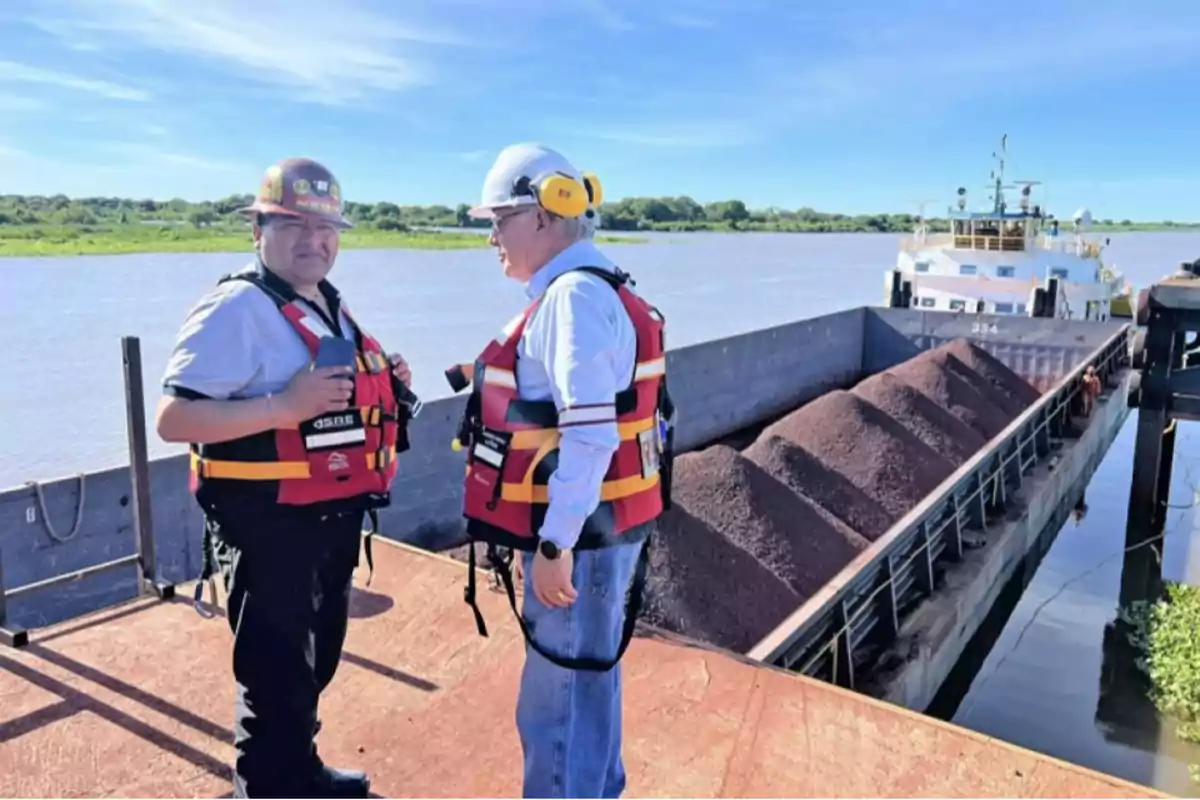 Dos personas con chalecos salvavidas y cascos de seguridad están de pie junto a un barco cargado de material en un río.