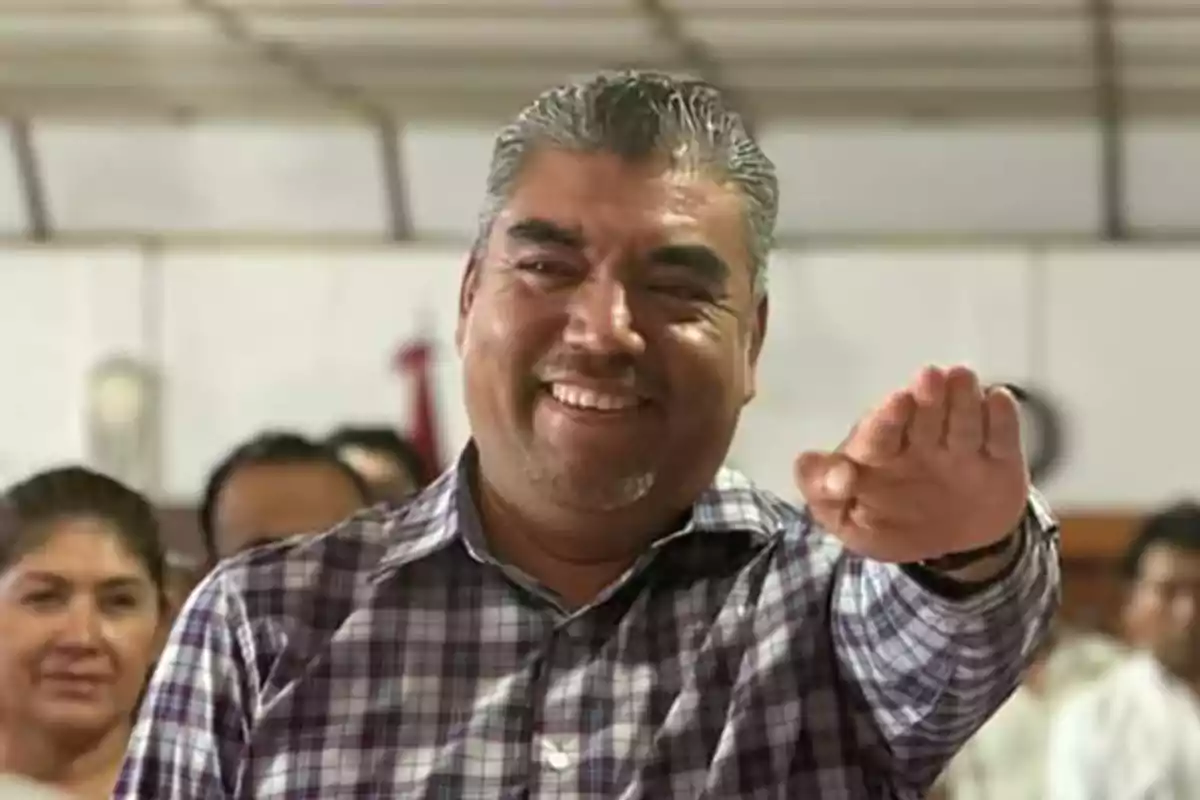 Hombre de cabello canoso y camisa a cuadros sonríe mientras extiende el brazo derecho al frente con la palma hacia abajo en un entorno interior con varias personas al fondo Hombre de cabello canoso y camisa a cuadros sonríe mientras extiende el brazo derecho al frente con la palma hacia abajo en un entorno interior con varias personas al fondo