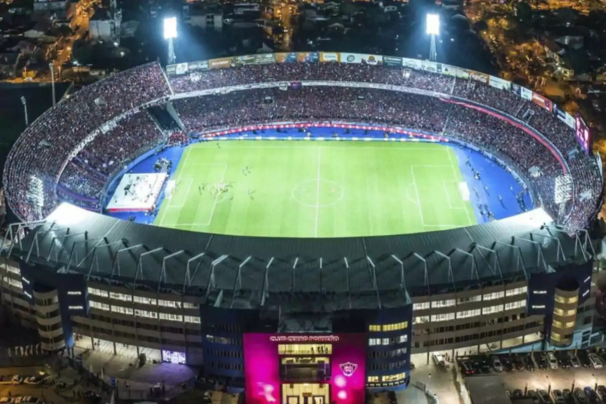 Vista aérea de un estadio de fútbol iluminado durante un partido nocturno con gradas llenas de espectadores y un campo de juego verde.