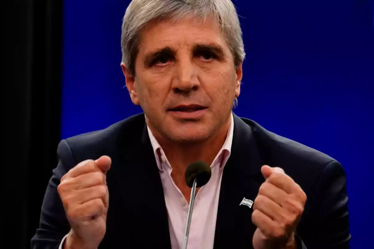 Un hombre de cabello canoso y traje oscuro gesticula con las manos frente a un micrófono sobre un fondo azul.