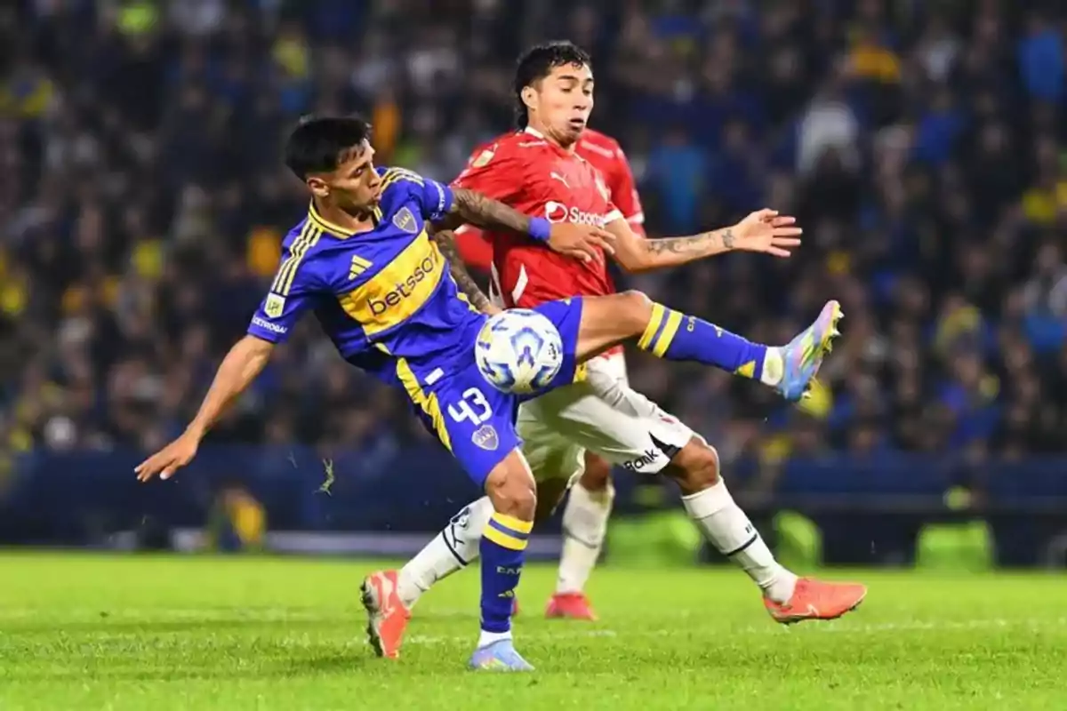 Dos jugadores de fútbol compiten por el balón en un partido, uno con uniforme azul y amarillo y el otro con uniforme rojo y blanco, en un estadio lleno de espectadores. Dos jugadores de fútbol compiten por el balón en un partido, uno con uniforme azul y amarillo y el otro con uniforme rojo y blanco, en un estadio lleno de espectadores.