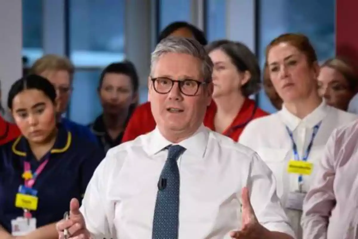 Un hombre con gafas y corbata habla frente a un grupo de personas, algunas de las cuales llevan uniformes médicos.