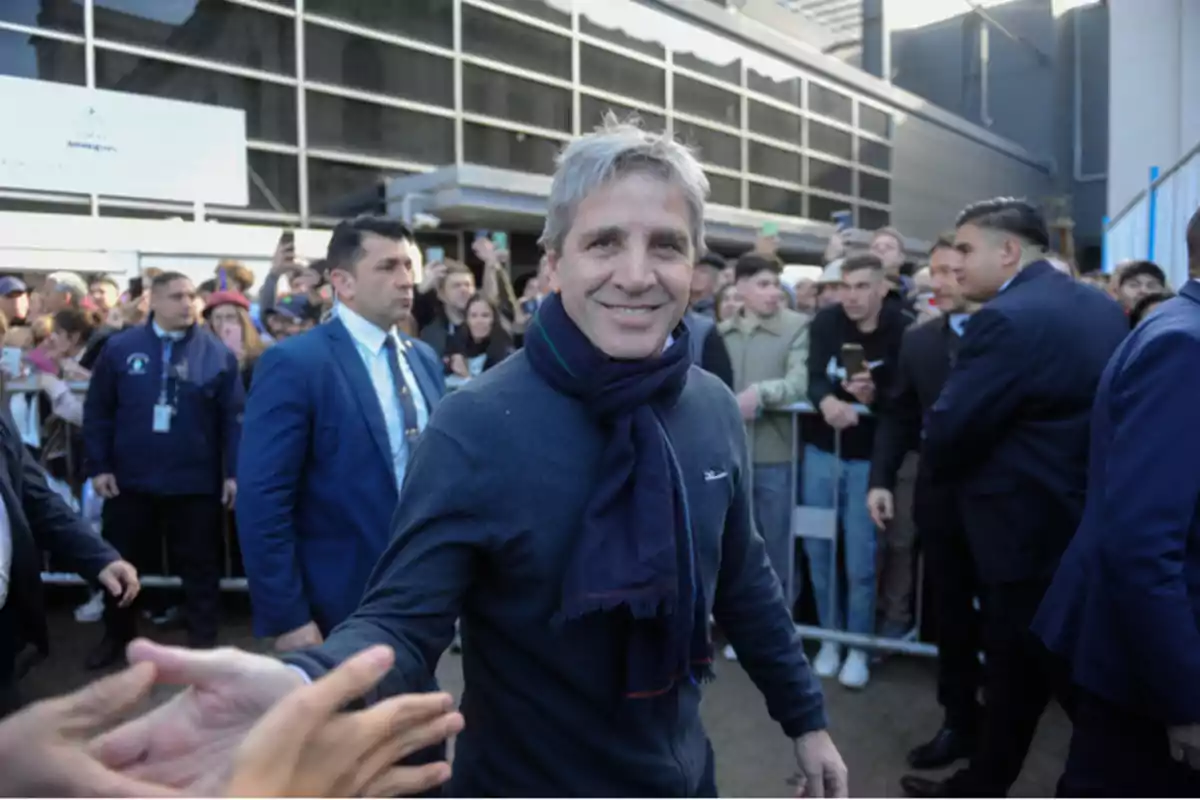 Un hombre sonriente con bufanda azul saluda a una multitud mientras es escoltado por personal de seguridad.