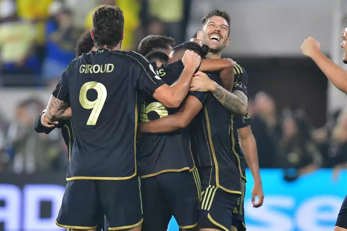 Jugadores de fútbol con uniforme negro celebran abrazados en la cancha mientras uno de ellos sonríe ampliamente Jugadores de fútbol con uniforme negro celebran abrazados en la cancha mientras uno de ellos sonríe ampliamente