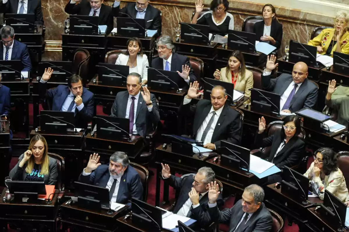 Un grupo de personas en una sesión parlamentaria levantando la mano para votar.