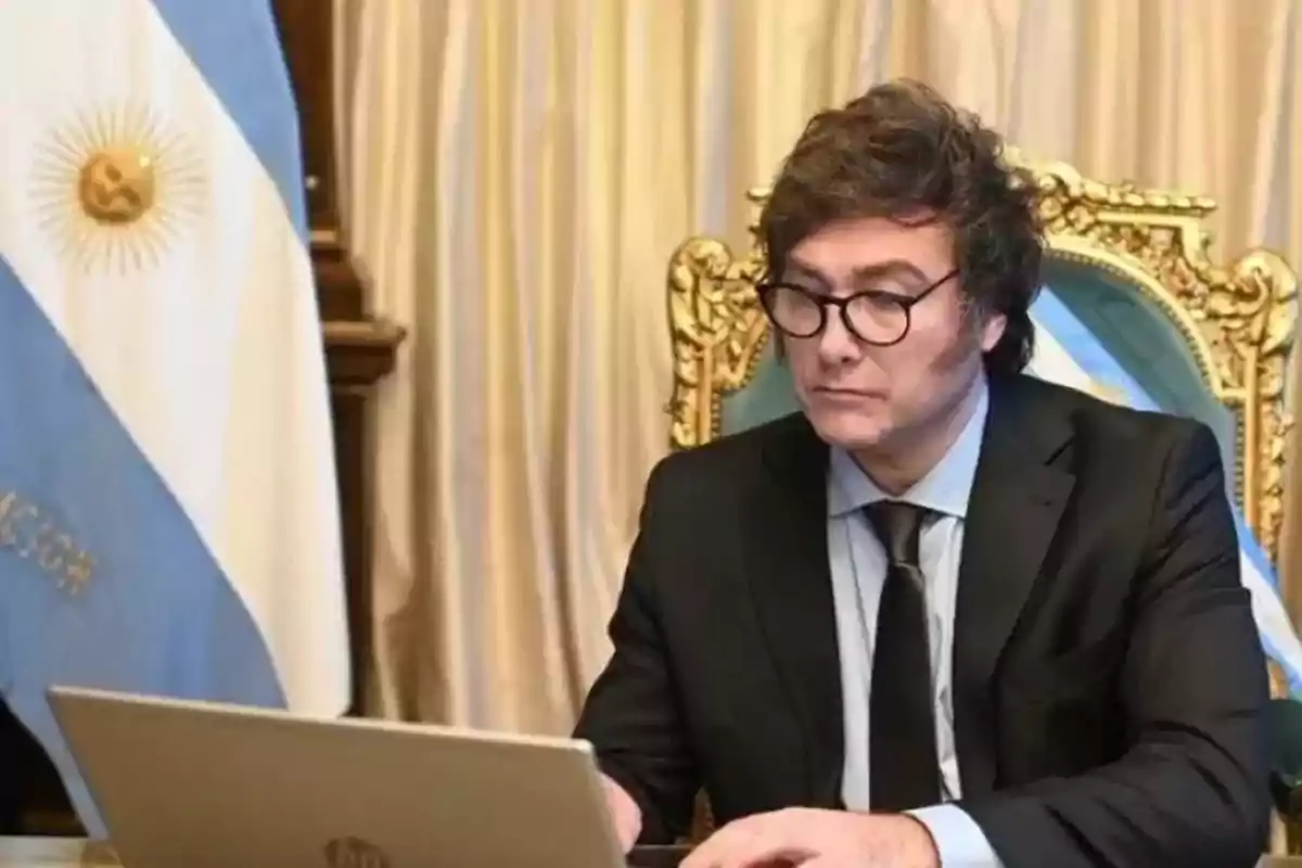 Un hombre con gafas y traje oscuro está sentado frente a una computadora portátil en una sala con cortinas doradas y una bandera argentina.