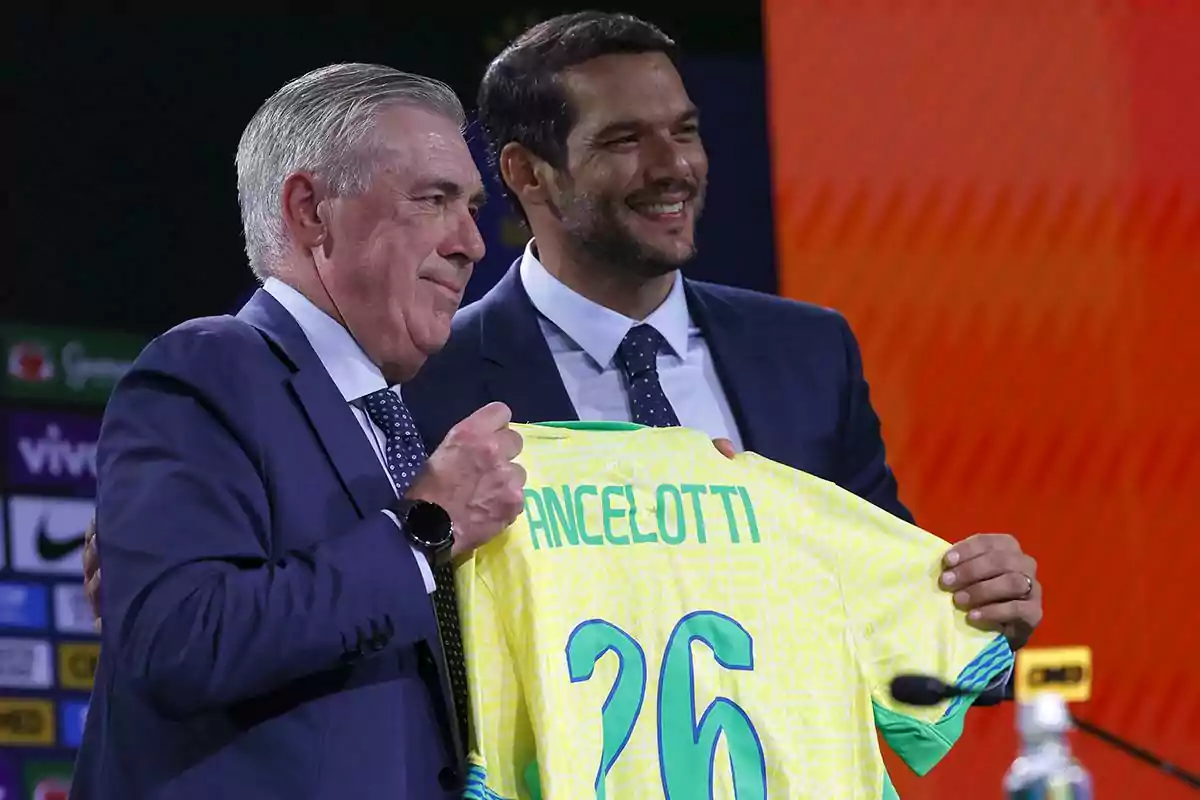 Dos hombres con traje sonríen mientras sostienen una camiseta de fútbol amarilla con el nombre Ancelotti y el número 26