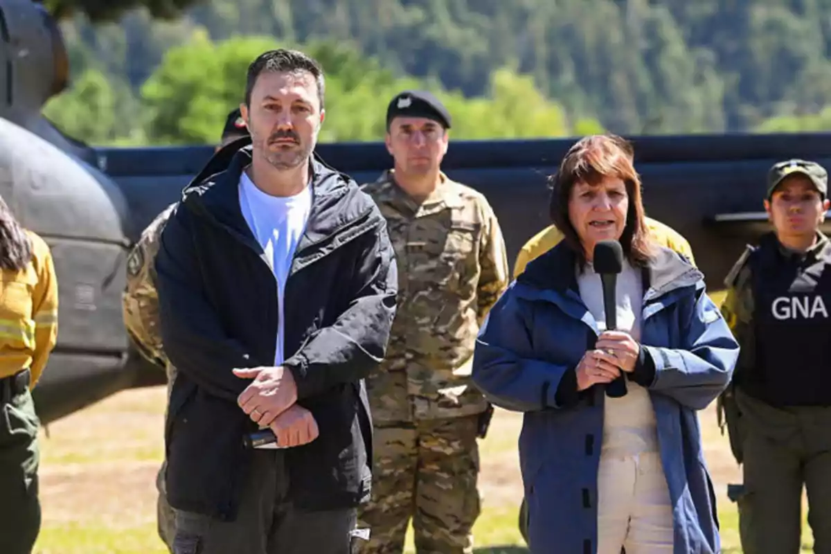 Un grupo de personas, algunas en uniforme militar, están de pie al aire libre; una mujer sostiene un micrófono mientras habla.