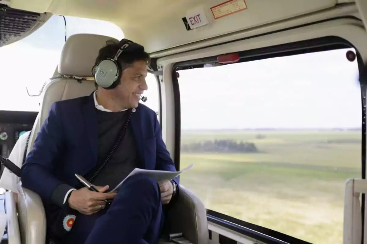 Un hombre con auriculares y un micrófono está sentado en un helicóptero, mirando por la ventana mientras sostiene un bolígrafo y un documento.