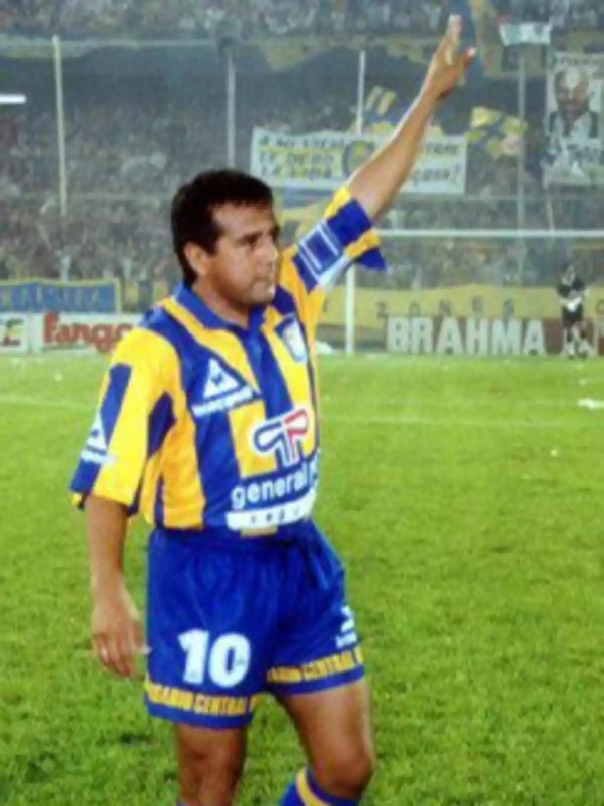 Jugador de fútbol en uniforme azul y amarillo levantando el brazo en un estadio lleno de espectadores.