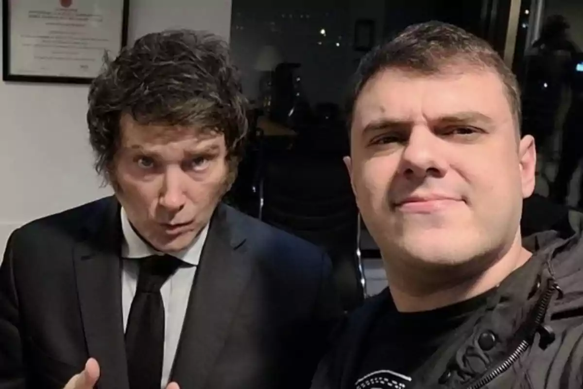 Dos personas posan para una selfie en un ambiente interior, una de ellas lleva traje y corbata mientras que la otra viste una chaqueta casual.