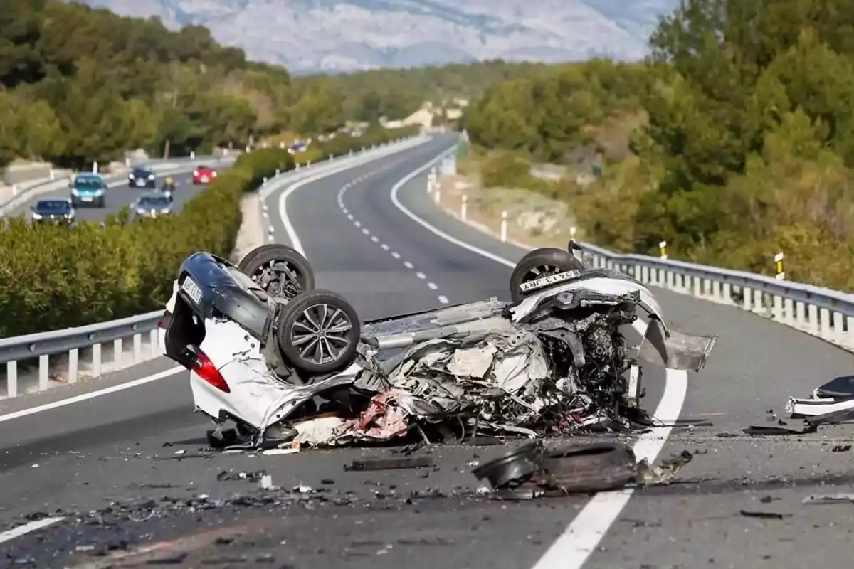 Auto volcado y destruido en medio de una carretera con escombros esparcidos y otros vehículos al fondo